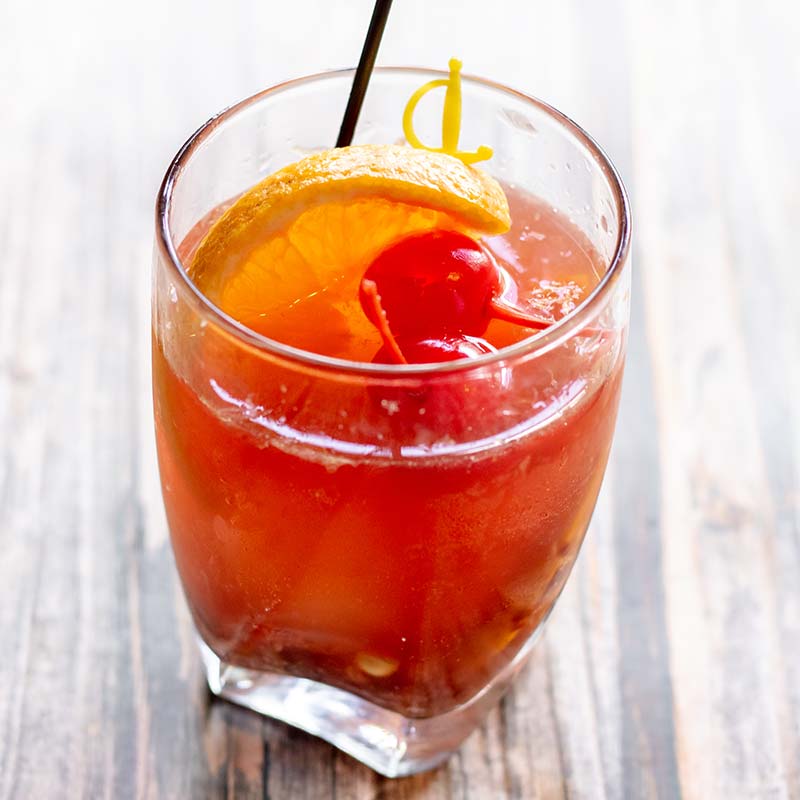 Red cocktail with orange slice, cherry, and straw at a restaurant near Amnicon Falls on wood table.